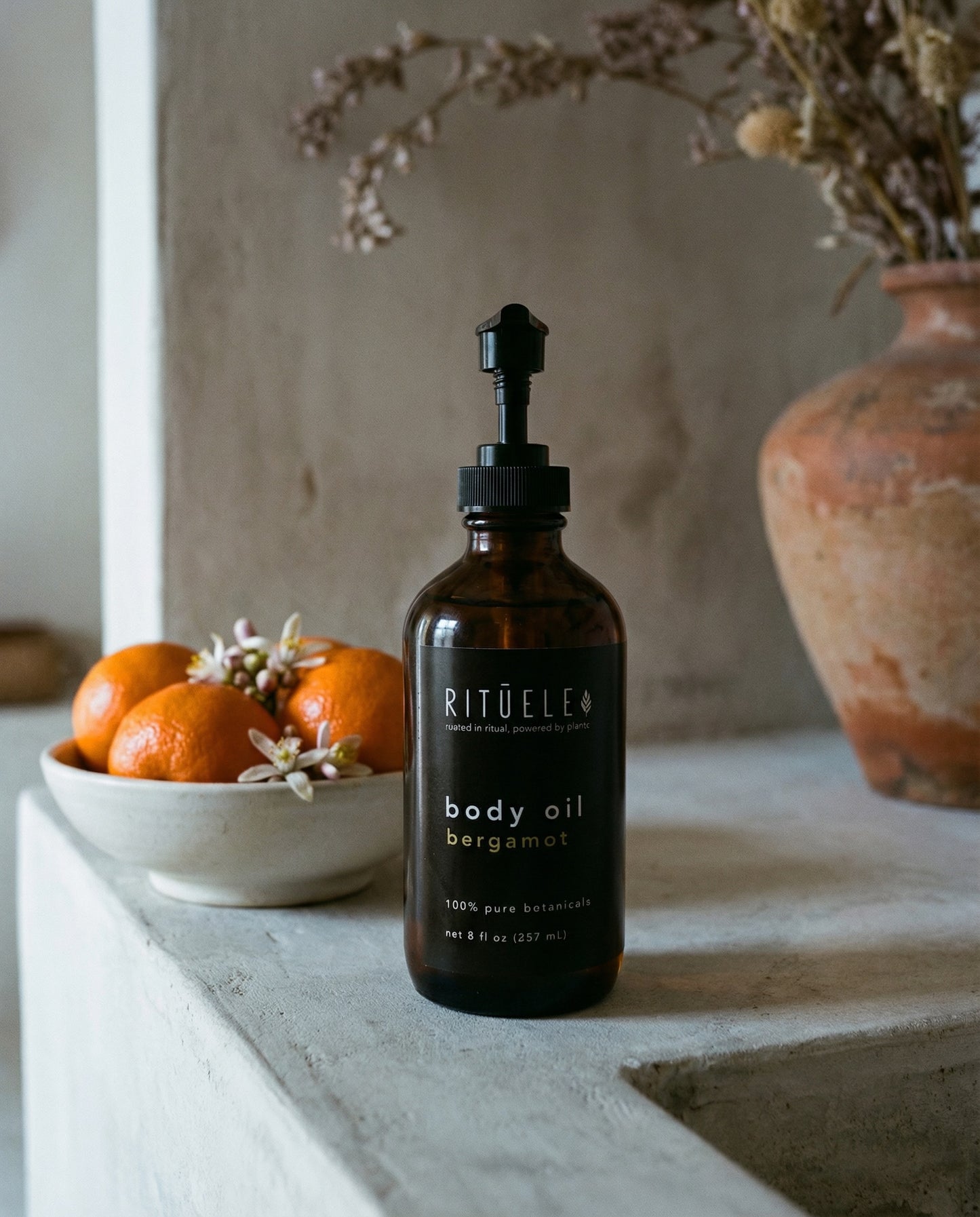 Rituelle body oil bottle on a surface with oranges and a vase in the background